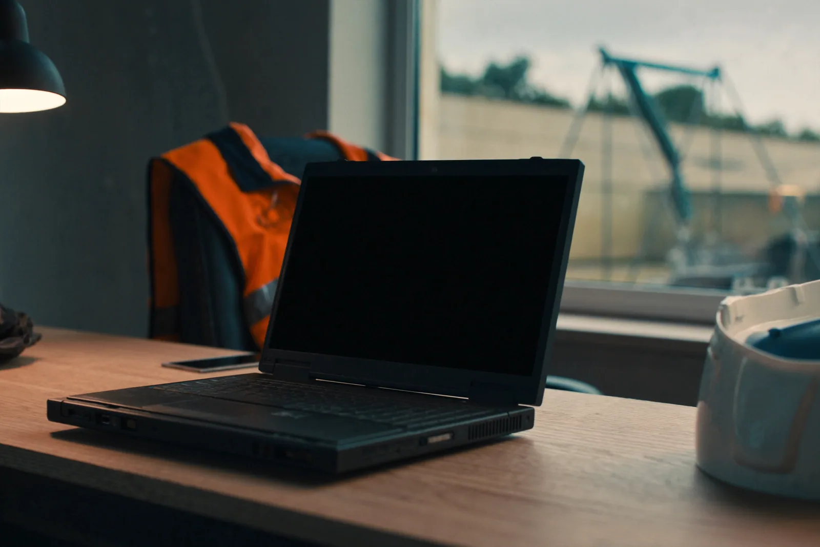 A rugged laptop with its screen off on a site office desk, a hi-vis vest on the chair and a crane through the window
