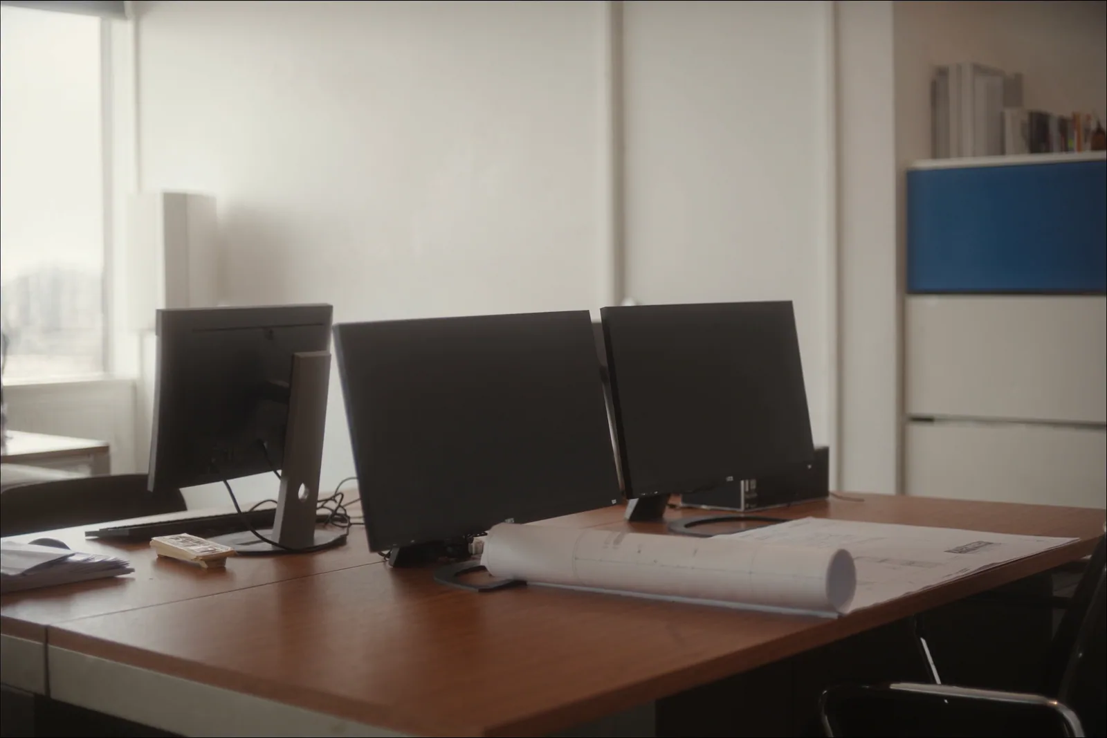 Two dark monitors and a rolled plan on a timber desk in a bright professional office
