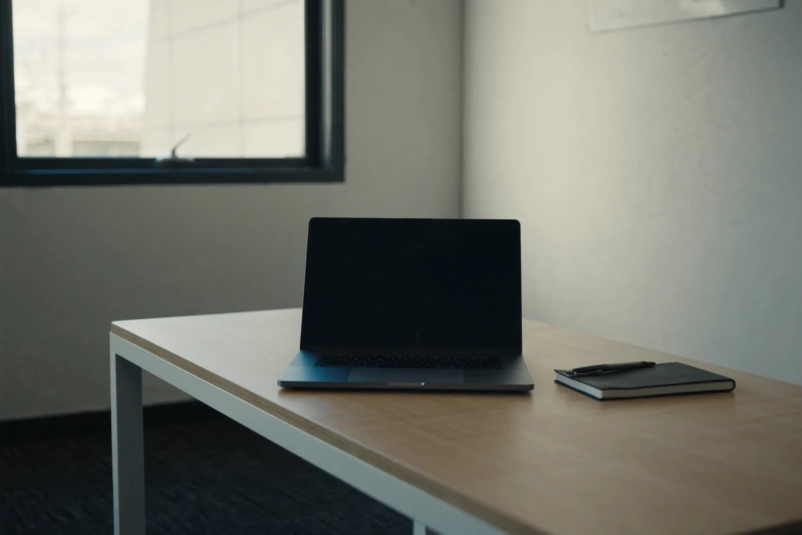 A laptop with its screen off and a notebook on a timber desk by a window, ready for a review