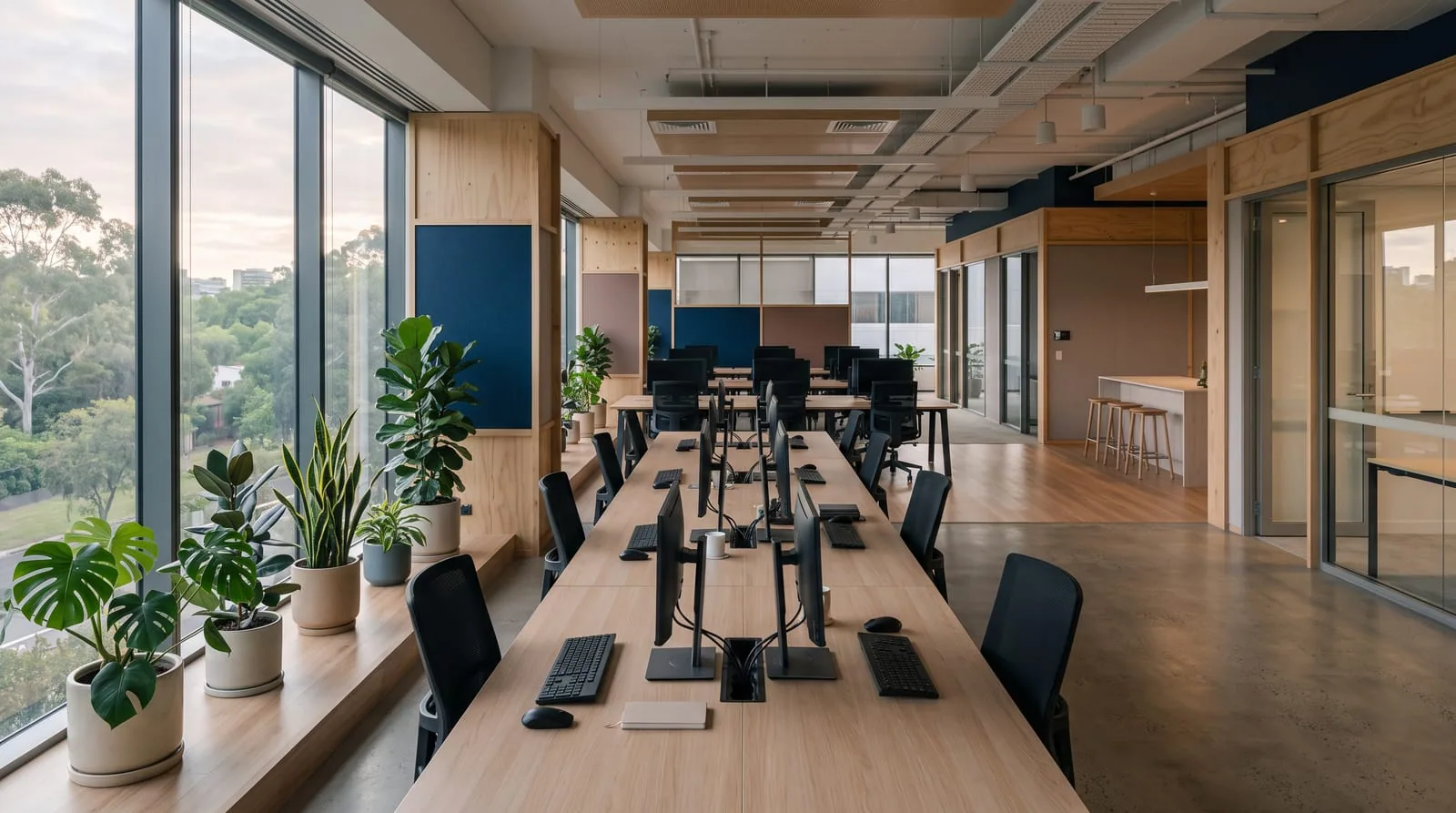 An open-plan office with timber desks and monitors, daylight through tall windows
