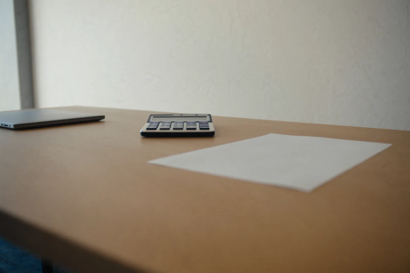 A closed laptop, a calculator and a blank sheet of paper on a timber desk